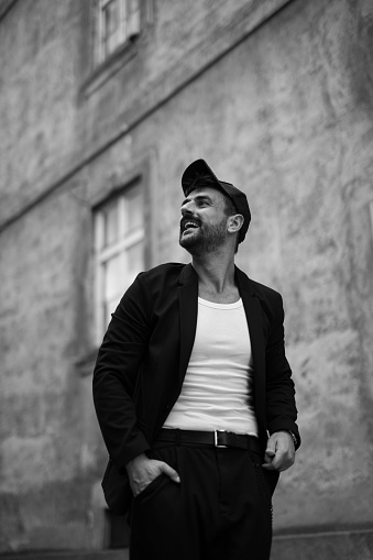 Spontaneous black and white image of a cool looking, bearded, young, stylishly dressed man, wearing a suit jacket and sports hat, standing outdoors, facing up, smiling. Street style enjoyment, with his hands in a pocket, stylish concept