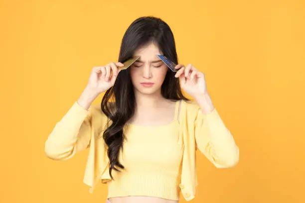 Photo of Asian woman close her eyes while holding credit card with feeling stressed, broke, angry and frustrated on yellow background.