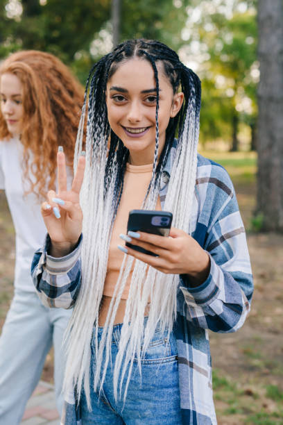 istock Smiling at camera young beautiful woman with African pigtails using 4g wireless connection on mobile phone for updating profile online. Millennial best friends using smart phone. Technology and friendship concept 1422701519