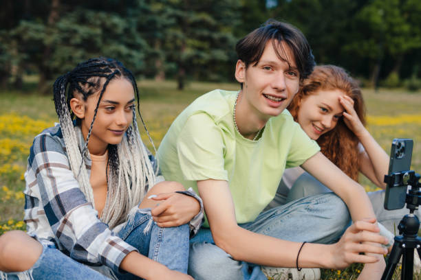 istock A group of three people using a tripod and phone to take a group photo while they sitting on the grass in the summer park. Community concept with multicultural and multiethnic friends. Friendship, photo, selfie, communication concept. 1422701532