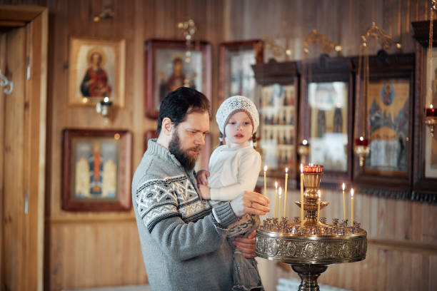 A Russian man with a beard and a daughter is standing in an Orthodox Church, lighting a candle and praying in front of the icon. stock photo