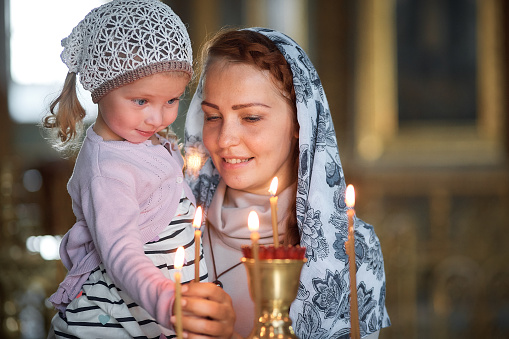 Russian beautiful woman in a scarf and with red hair holding a little girl and lights a candle in front of an icon in the Russian Orthodox Church.