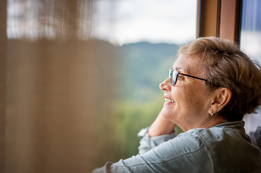Portrait of a beautiful mature woman in glasses standing near the window in profile