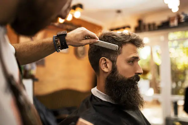 Photo of Young barber making haircut to a bearded man using comb