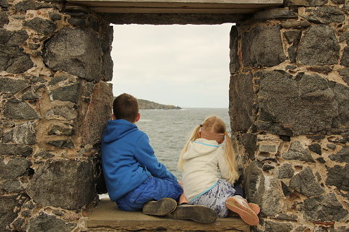 Young boy and girl sitting in the ruined window of a building facing away out to sea on a grey overcast day. They are wearing warm clothing