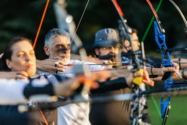 Women and men practicing archery training with recurve bow on open field before sunset.