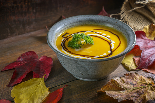 Autumn soup from red kuri squash with parsley garnish in a rustic bowl on a dark wooden table with colorful leaves for Thanksgiving or Halloween, copy space, selected focus, narrow depth of field