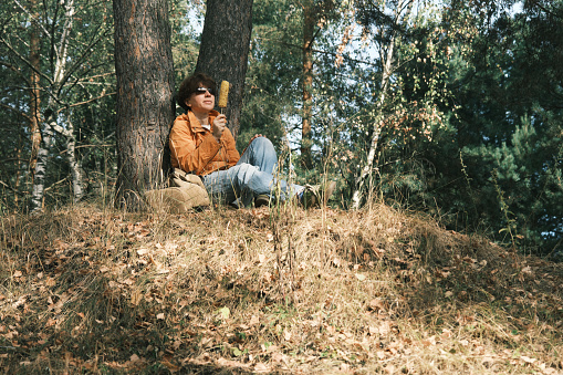 Cheerful mature woman is enjoying grilled corn while resting in autumn nature park