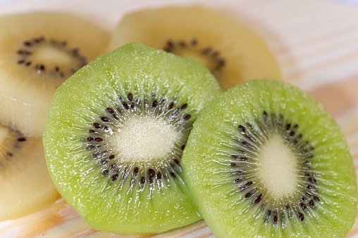 Stock photo showing close-up, elevated view of a layer of kiwi fruit slices on a wood grain background. Pieces of Chinese gooseberry displaying fuzzy brown skin and bright green flesh with ring of black seeds, healthy eating poster wallpaper background design.