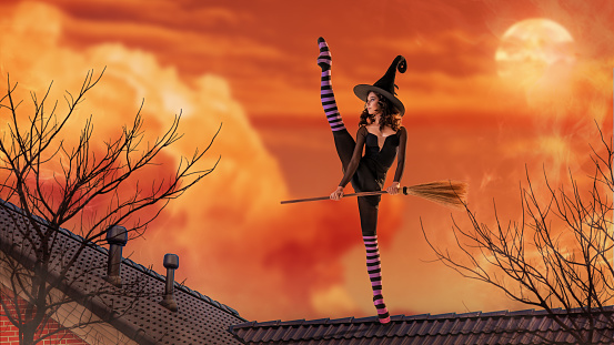 ballerina in pointe shoes in a black witch costume with a hat and a broom is dancing on the roof of old house against the backdrop of ominous bright sunset.