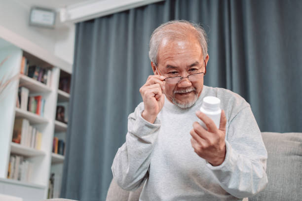 丸薬を食べているアジアの高齢男性患者は、老人ホームケアで自己回復します。眼鏡をかけている老人は処方箋と内容を読んでいます。医療保険のコンセプト - senior adult nursing home eating home interior ストックフォトと画像
