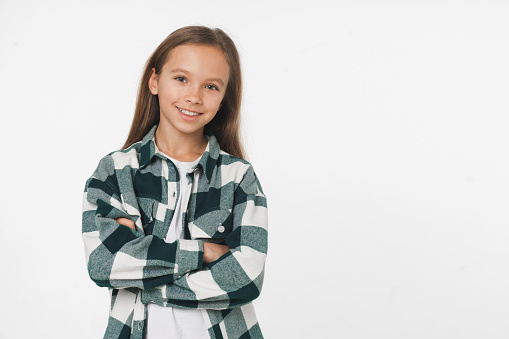 Happy smart young preteen girl schoolgirl in checkered shirt looking at camera smiling with toothy smile with arms crossed isolated in white background