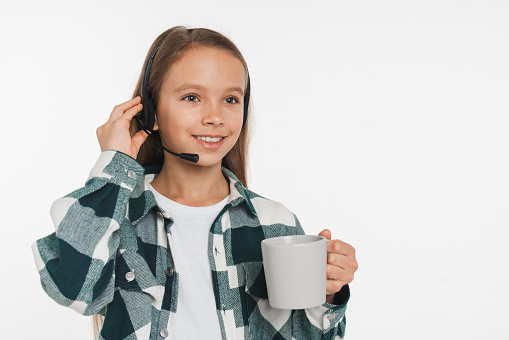 Young happy IT support manager customer support preteen girl teenager schoolgirl in headset drinking coffee tea isolated in white background