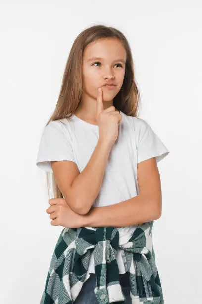 Photo of Vertical portrait of pensive thoughtful caucasian small schoolgirl in white t-shirt and checkered looking at camera thinking isolated in white background