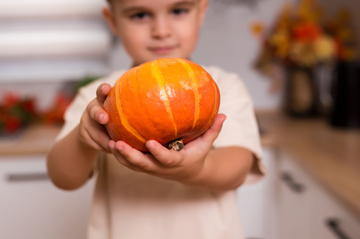 Caucasian little happy boy holds a fresh pumpkin at kitchen. Healthy lifestyle, diet concept, vegetarianism, raw food and autumn holidays.