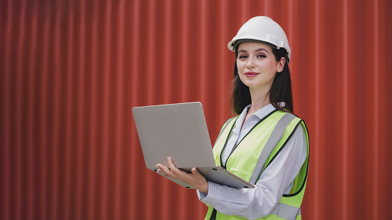 Portrait Caucasian woman with Container box Shipping Logistics Engineering of Import/Export Transportation Industry, Female Safety Transport Engineer holding laptop standing confidently at shipyard.