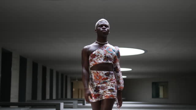 Low angle tracking shot of self assured Black person in stylish crop top and skirt putting hands on waist while walking towards camera inside underground building with dim illumination