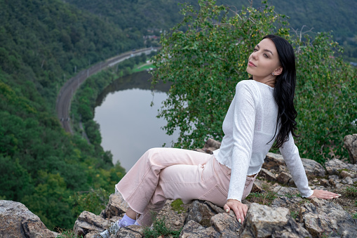 A young woman sits on a rock with a view of the forest and mountains and river vltava. Typical country landscape in europa