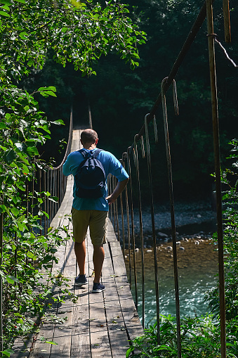 a male traveler with a backpack on his shoulders walks along a suspension bridge over a mountain river