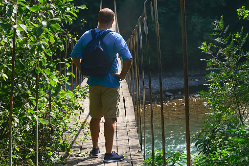 a male traveler with a backpack on his shoulders stands at the beginning of a suspension bridge over a mountain river and prepares to overcome it