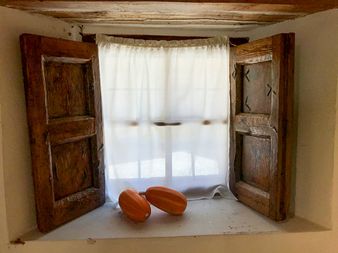window with rustic wooden doors, white fabric curtain and two natural pumpkins as decoration.