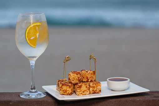 snack called Dadinho de tapioca made with rennet cheese and tapioca flour, served with pepper jelly. tropical drink and beach in the background.