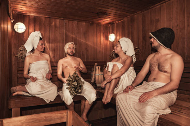 Russian bathhouse. Two couple relaxing and sweating in wooden sauna with hot steam. Four person with bath besoms resting on bench in spa complex. Wellness, self care, healthy concept. Copy text space casa de baños rusa. dos parejas relajándose y sudando en sauna de madera con vapor caliente. cuatro personas con besoms de baño descansando en el banco en el complejo de spa. bienestar, autocuidado, concepto saludable. copiar espacio de texto