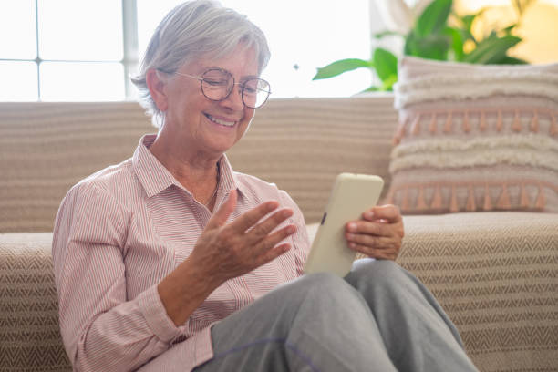 행복한 유쾌한 노인 여성은 스마트 폰을 손에 들고 바닥에 앉아 휴식을 취합니다. 웃는 성숙한 아가씨가 소셜 네트워크에서 채팅하고, 재미있는 비디오를보고, 집에서 모바일 응용 프로그램을 � - telephone window mobile phone addiction 뉴스 사진 이미지