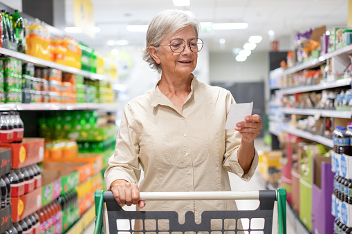 Caucasian elderly woman checking her list while purchasing household products in supermarket. Senior female with shopping cart in departement store