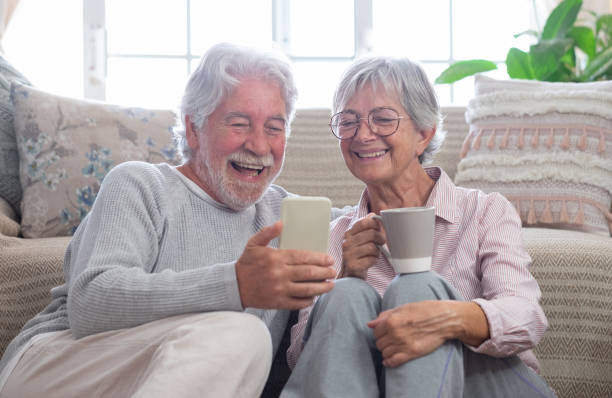un couple de personnes âgées souriant et agréable se détend à la maison assis par terre en regardant ensemble des vidéos amusantes sur un téléphone portable. - telephone window mobile phone addiction photos et images de collection