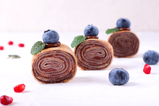 Rolled crepes with chocolate filling topped with blueberry and mint on white table. Served dessert
