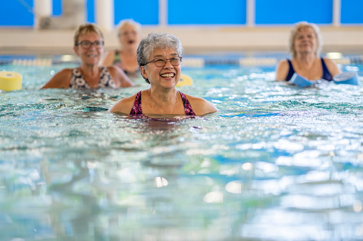 A senior woman is seen swimming in a pool as she laughs and enjoys the activity.  She is wearing a swim suit and is among friends as she participates in an Aqua fitness class.