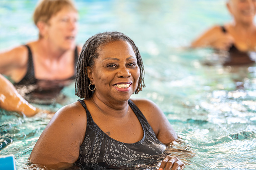 A small group of seniors are seen swimming in a pool as they participate in an Aqua fitness class together.  They are each wearing swim suits and have smiles on their faces as they enjoy the activity.