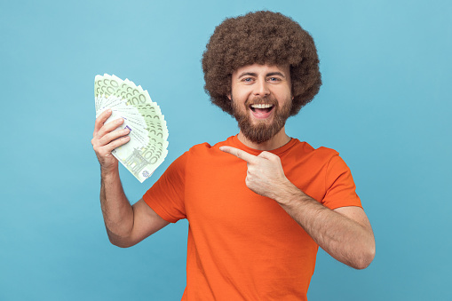 Portrait of delighted optimistic man with Afro hairstyle wearing orange T-shirt holding euro banknotes, being happy to win lottery. Indoor studio shot isolated on blue background.