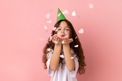 Portrait of little girl wearing white T-shirt and party hat blowing heart shape glitter, enjoying birthday, celebrating event, festive mood. Indoor studio shot isolated on pink background.