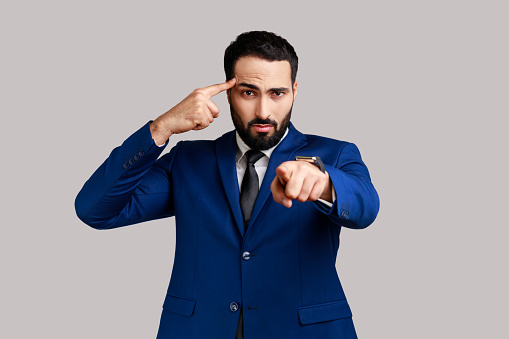Displeased bearded man holding finger near temple and pointing on you, showing stupid gesture, mocking, wearing official style suit. Indoor studio shot isolated on gray background.
