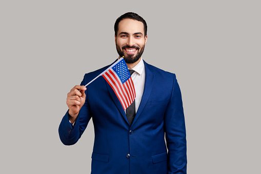 Portrait of positive optimistic bearded man holding united states of america flag, patriotism, independence, wearing official style suit. Indoor studio shot isolated on gray background.
