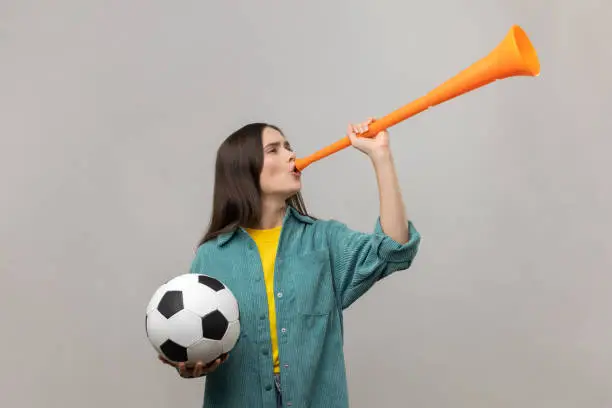 Photo of Excited woman blowing in horn holding soccer ball, celebrating victory of favourite football team.