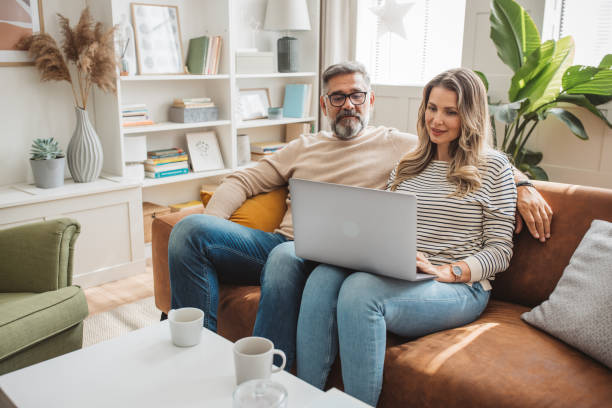 Shopping from home Middle aged couple sitting on sofa and using laptop. 55 59 years stock pictures, royalty-free photos & images