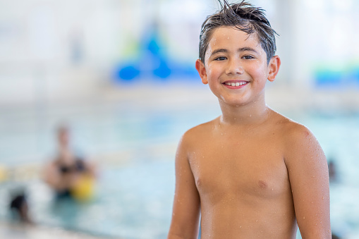 A young boy stands beside an indoor pool as he poses for a portrait.  He is smiling and wearing a swim suit as he prepares to participate in a swimming lesson.