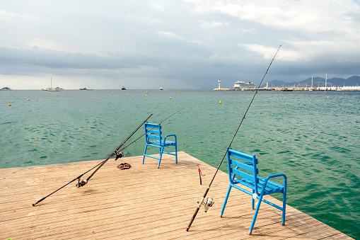 Fishing rods (spinnings) without people leaning against the chairs and the view from the pier on the Mediterranean Sea - the blue water of Cannes and a large ocean cruise liner and a lighthouse in the distance!