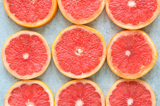 Stock photo showing citrus fruit slices on mottled grey background, modern minimalist photo of circular sliced pink grapefruit citrus fruit showing segments, seeds / pips and rind around edge, healthy eating concept photo for vitamin C and fruit juice.