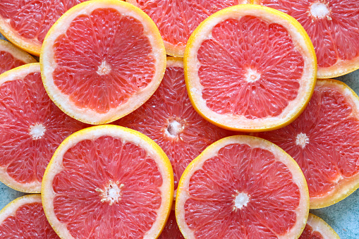 Stock photo showing citrus fruit slices on mottled grey background, modern minimalist photo of circular sliced pink grapefruit citrus fruit showing segments, seeds / pips and rind around edge, healthy eating concept photo for vitamin C and fruit juice.