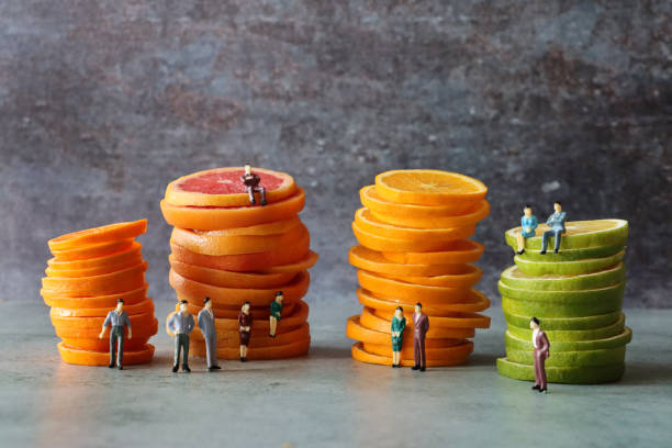 istock Image of rows of sliced citrus fruit piles surrounded by plastic model people, pink grapefruit slices stack, towers of orange slices, pile of chopped lime, mottled grey background, focus on foreground 1422869909