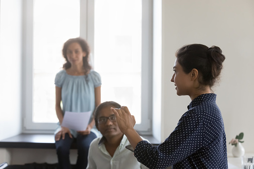 Young Indian mentor training diverse group of interns in office. Millennial business woman talking to team of employees in corporate meeting, Side view. Female leadership concept