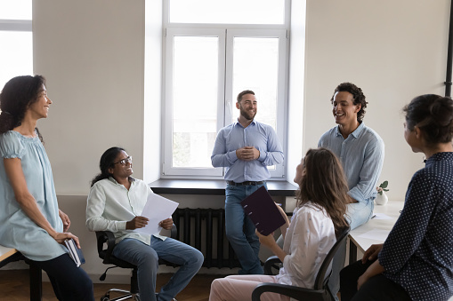 Cheerful coworkers meeting in open office space, negotiating on project tasks, collaborating on ideas, discussing decision, brainstorming, talking,. Business team sitting in circle, laughing
