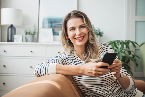 Mature woman at home relaxing on sofa. She is using smart phone.