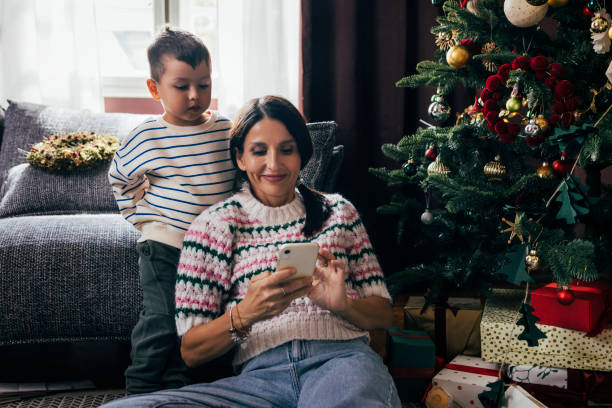 glückliche frau, die ihr telefon benutzt, während sie mit ihrem sohn am weihnachtsbaum sitzt - christmas shopping internet family stock-fotos und bilder