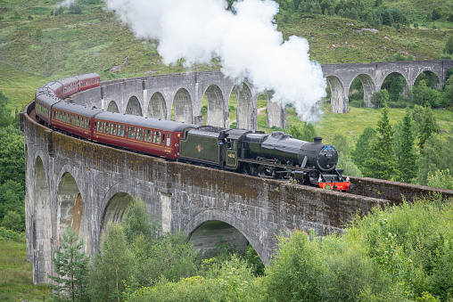Glenfinnan,Inverness-shire,Scottish Highlands-July 21 2022:The train driver waves and lets off steam to onlookers,as the iconic train,shown in Harry Potter films,crosses the tall viaduct,mid summer.