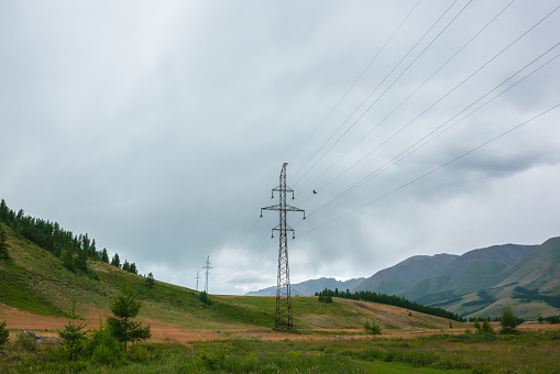 Dramatic mountain landscape with tall tower of power lines under cloudy sky. Atmospheric alpine scenery with electric tower with wires against high mountains in overcast. Electricity in mountains.